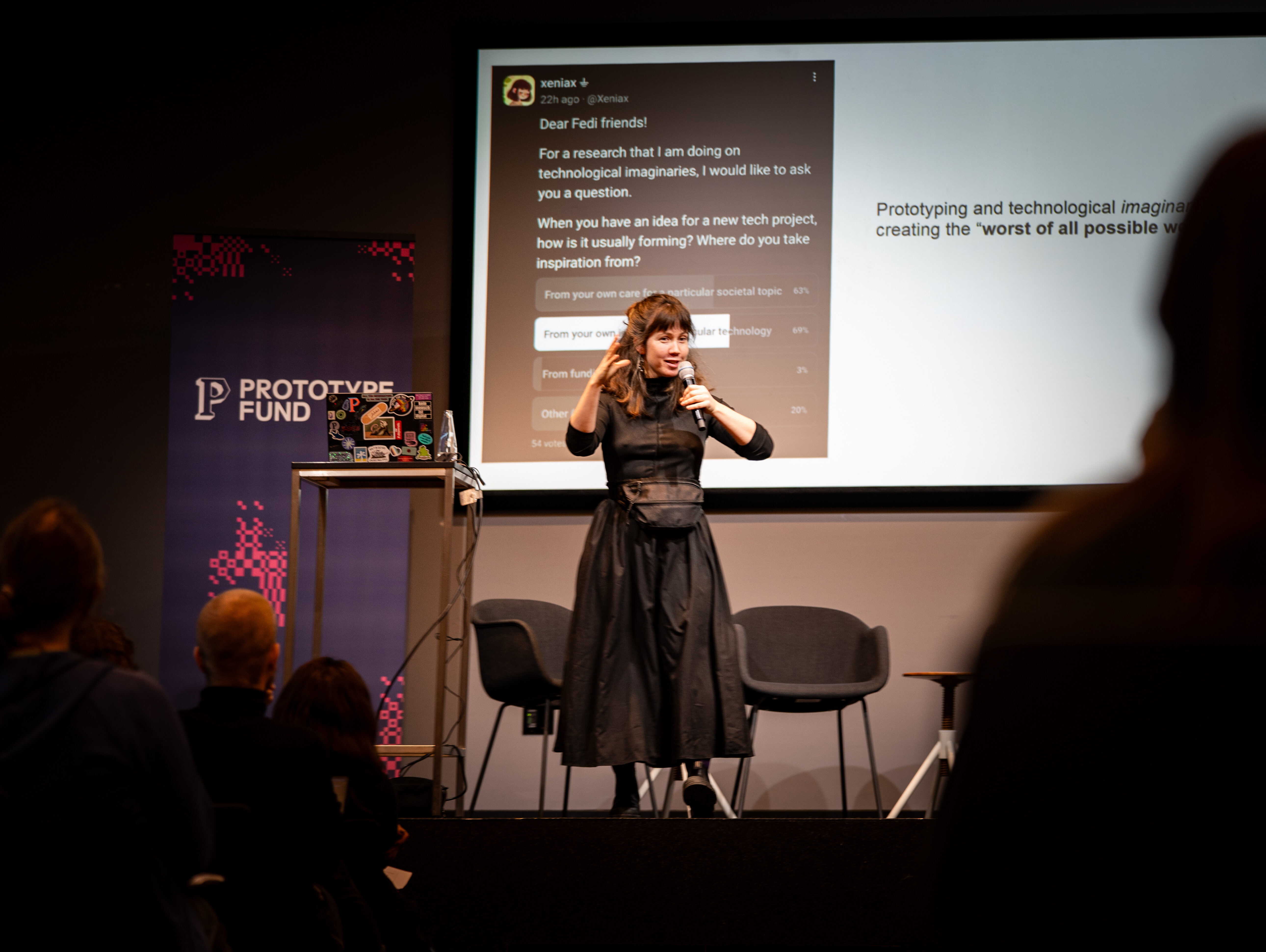 Ksenia steht auf der Bühne und redet in ein Mikrofon. Hinter ihr ein Roll-Up mit Prototype Fund Logo