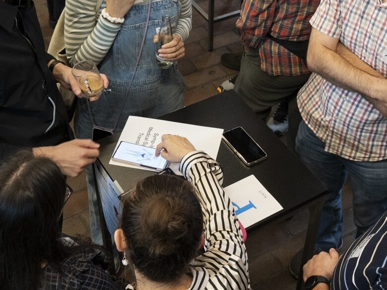 Foto von Personen, die um einen Tisch herum stehen. Darauf liegt eine Broschüre mit Prototype Fund P-Logo und ein Handy, auf dass eine der Personen zeigt.