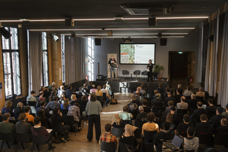 In einem Auditorium hört das Publikum einen Vortrag auf der Bühne.