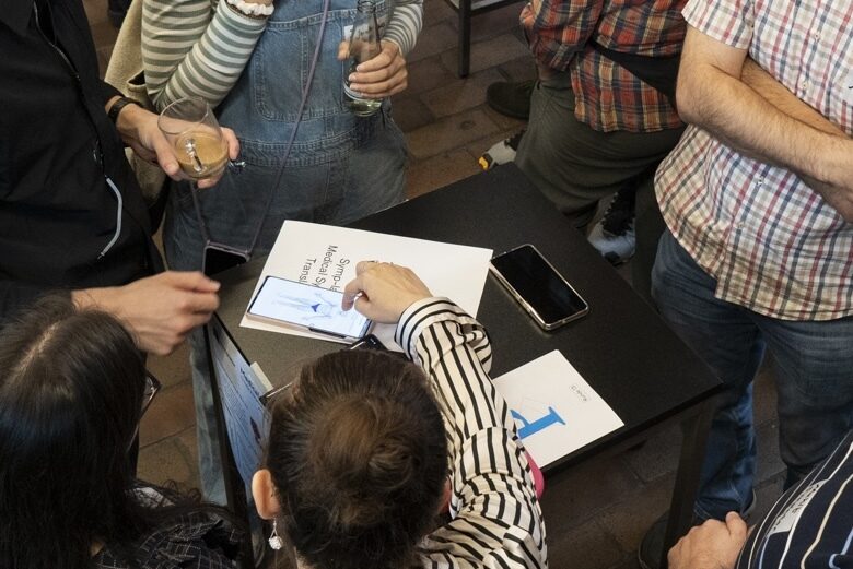 Foto von Personen, die um einen Tisch herum stehen. Darauf liegt eine Broschüre mit Prototype Fund P-Logo und ein Handy, auf dass eine der Personen zeigt.