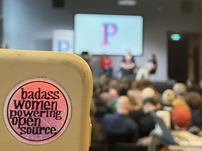 Sticker auf einem Handy: "badass women powering open source", im Hintergrund unscharf eine Bühne und ein Prototype Fund Logo auf einem Beamer