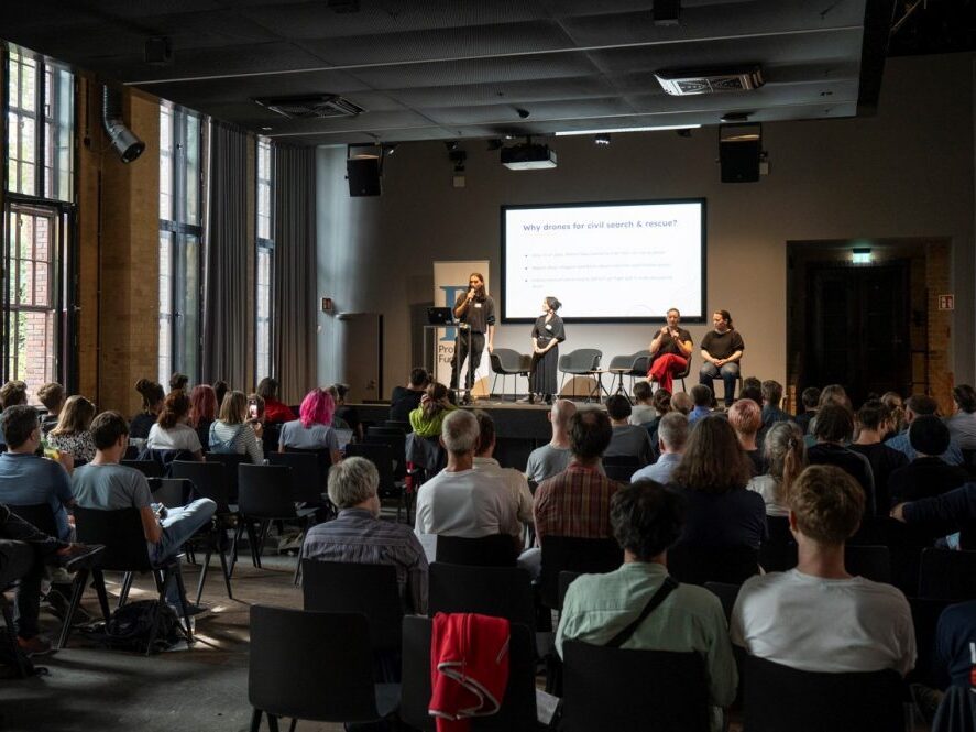 Vortrag des Projekts SearchWing beim Demo Day. Die zwei Personen aus dem Team stehen auf der Bühne, im Vordergrund ist das Publikum in Rückansicht zu sehen.