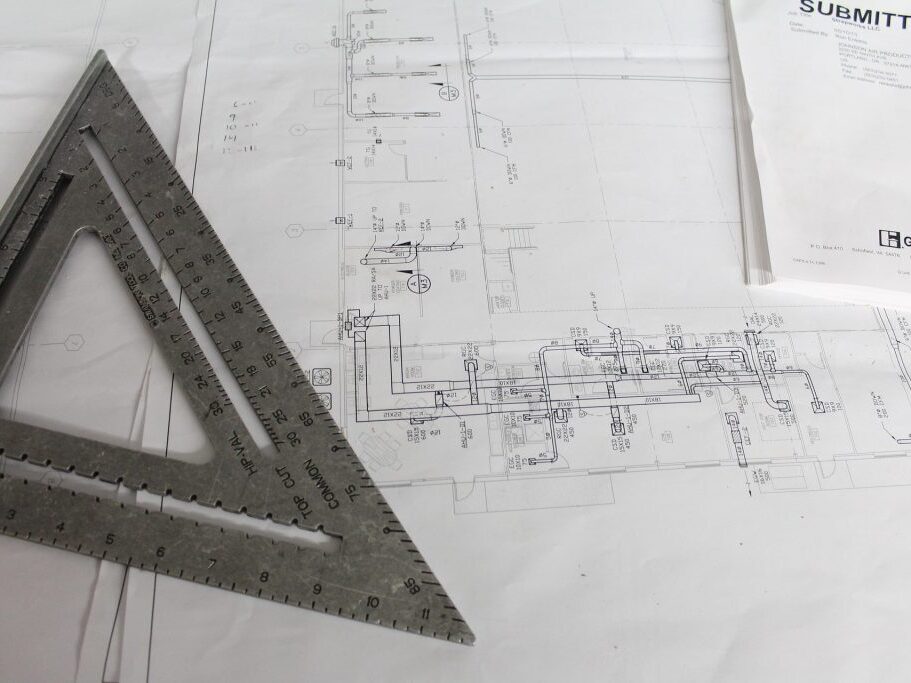 Foto eines Bauplans auf einem Tisch, darauf ein dreieckiges Lineal aus Metall