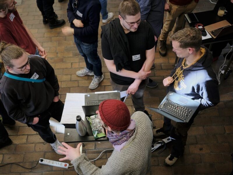 Eine Gruppe junger Menschen steht um ein kleines Podest im Rahmen des Demo Day. Einer hält einen Laptop aufgeklappt auf dem Unterarm. Je zwei von ihnen sind in ein Gespräch miteinander vertieft.