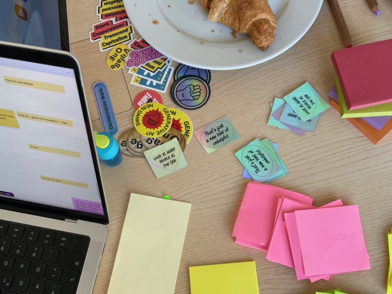 Foto eines Laptops auf einem Tisch, auf dem Sticker und Post-Its sowie ein Croissant auf einem Teller liegen.
