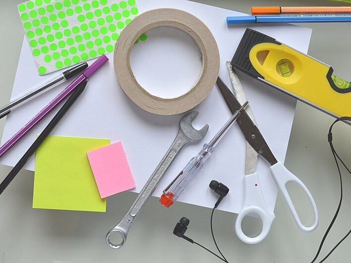 Eine Ansammlung von Gegenständen auf einer weißen Oberfläche. Darunter befinden sich weißes Papier, bunte Klebezettel, Klebeband, eine Schere, Stifte, eine Wasserwaage, Kopfhörer, ein Schraubenzieher und ein Ringratenschlüssel.