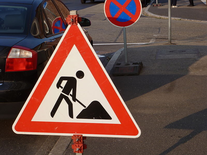 Foto eines Verkehrszeichen "Achtung Baustellen" im Hintergrund ein parkendes Auto sowie ein Halteverbotszeichen