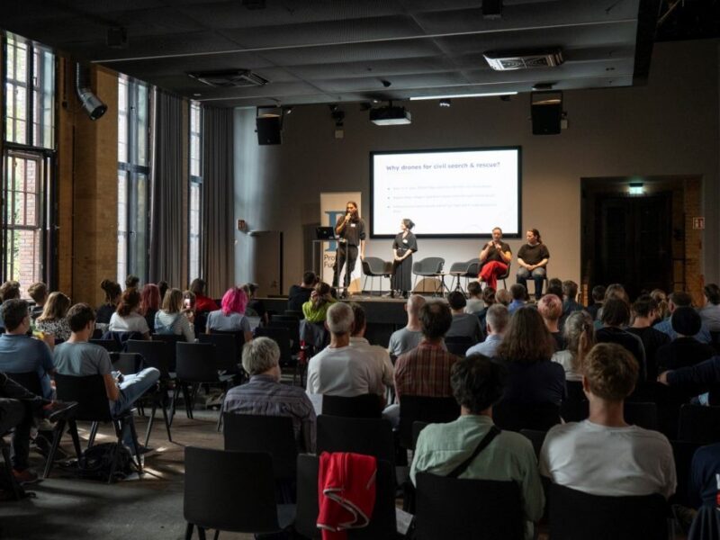 Vortrag des Projekts SearchWing beim Demo Day. Die zwei Personen aus dem Team stehen auf der Bühne, im Vordergrund ist das Publikum in Rückansicht zu sehen.