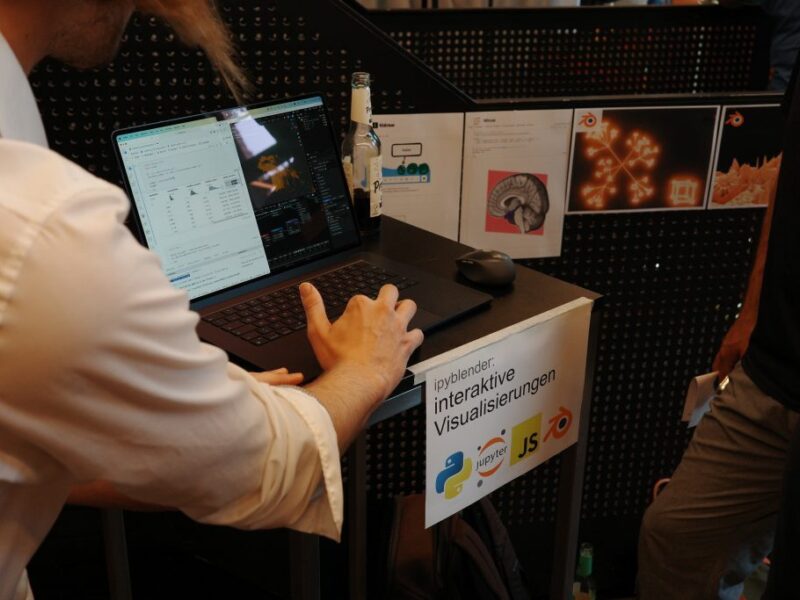 Foto: Ein Laptop steht auf einem Stehtisch, eine Hand zeigt etwas darauf. Ein Schild an dem Tisch sagt "ipyblender: interaktive Visualisierungen", im Hintergrund einige 3D-Graphiken