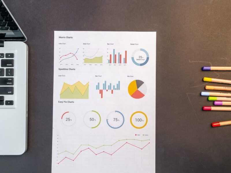 Bunte Stifte, ein Papier mit verschiedenen Diagrammen und ein aufgeklappter Laptop auf einem Tisch.