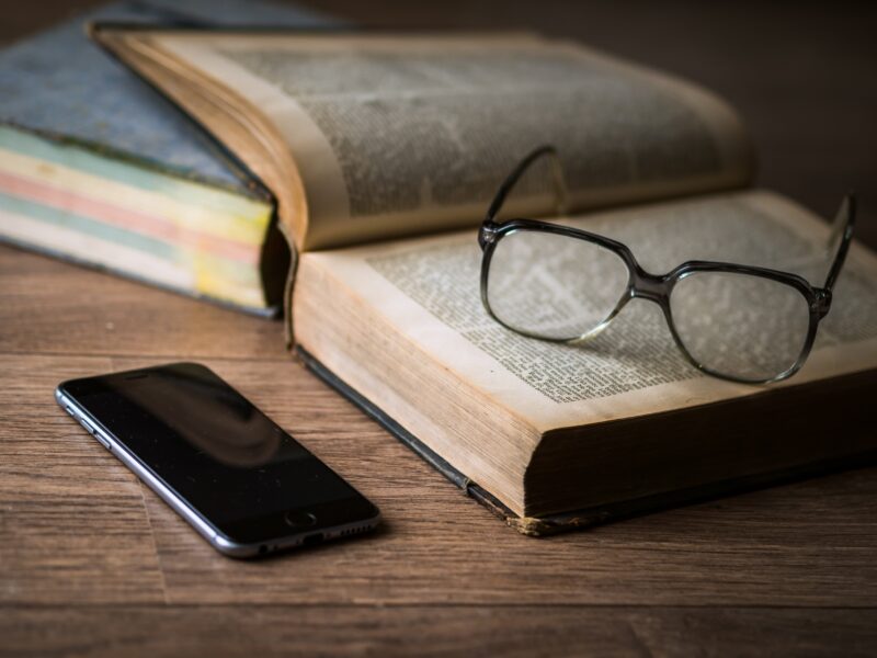 Ein Foto von einem aufgeschlagenen Buch, darauf liegt eine Brille. Daneben ein Smartphone.