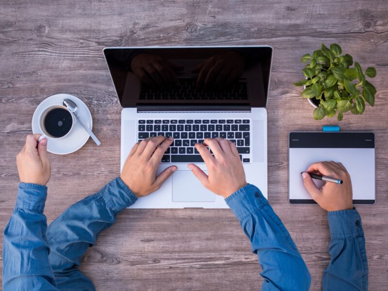Stockfoto: Vogelperspektive auf einen Schreibtisch, darauf vier Arme mit dem selben Hemdsärmel, die einerseits eine Kaffeetasse festhalten, auf einem Laptop tippen und auf einem Tablet zeichnen.