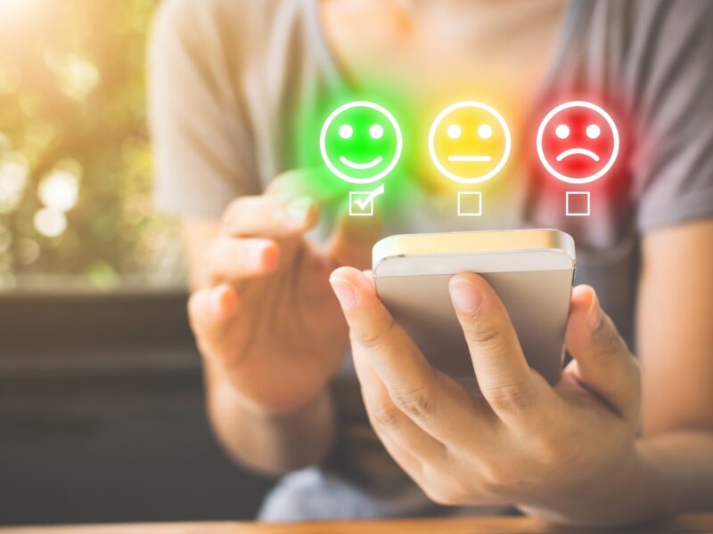 Foto von einer Hand, die ein Handy in der Hand hält. Darüber schweben neonfarbene Smileys: Ein lächelnder in grün, ein neutraler in gelb und einer mit heruntergezogenen Mundwinkeln in rot. Darunter sind Checkboxen, von denen die unter dem grünen Smiley ausgewählt ist.